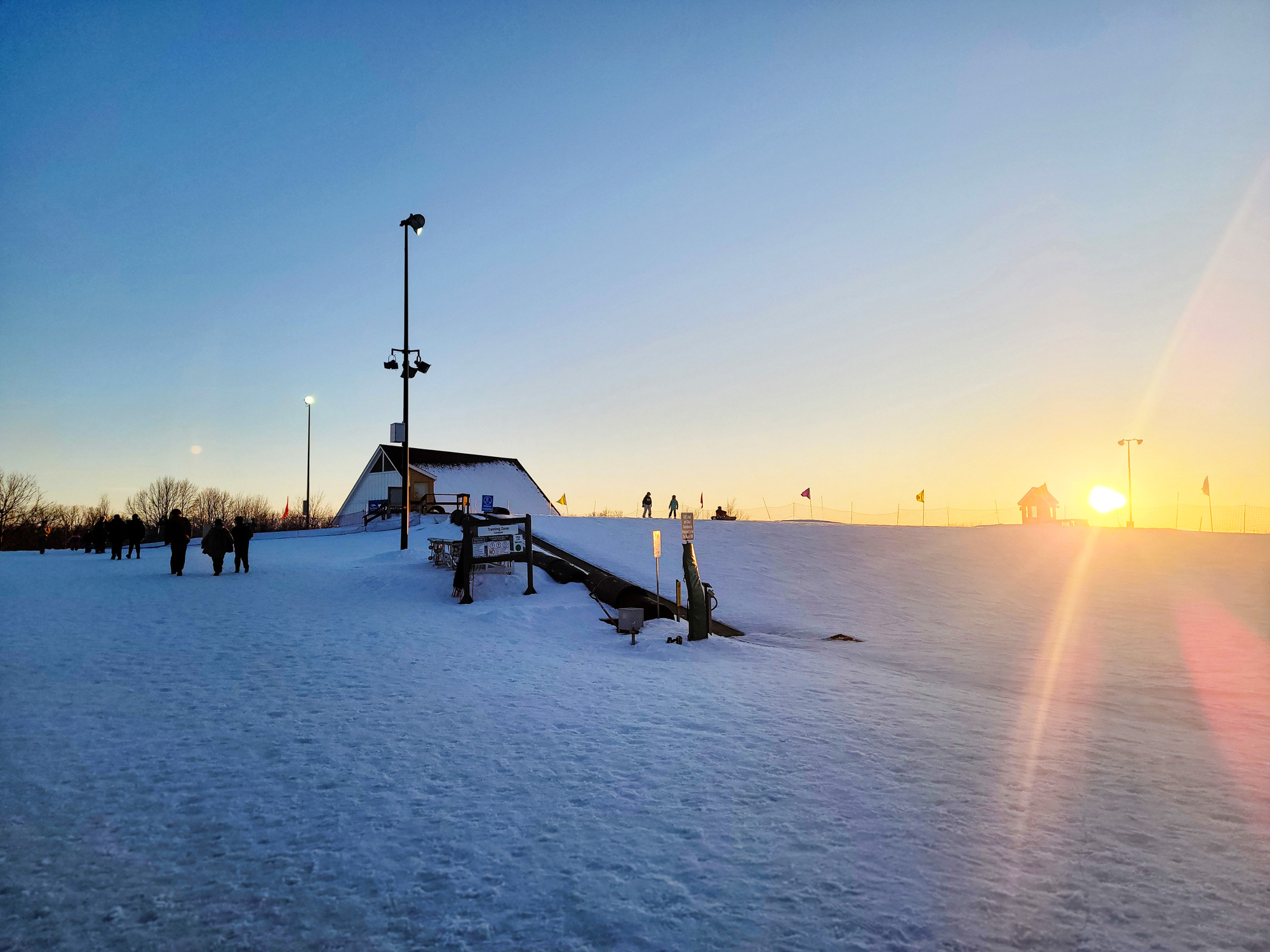 A landscape photograph of a snowy hill, there is a hut in the background and a jump in the middle ground. The sun is setting in the background