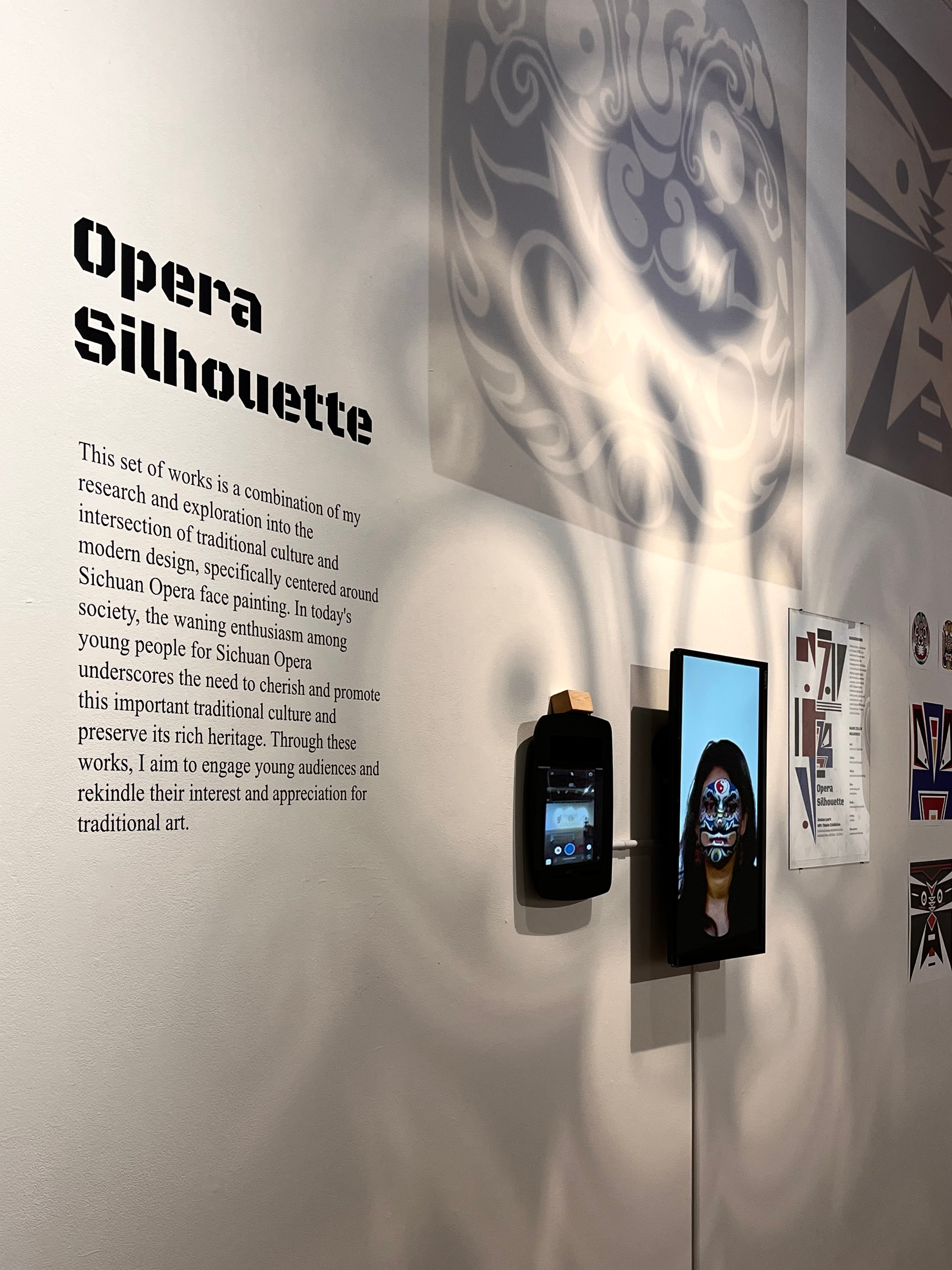 Image of Junlan's thesis exhibition work titled "Opera Silhouette." Includes vinyl didactic, tablet with interactive interface to take selfies with AR opera masks, and shadows from hanging prints of opera masks on transparent substrate.
