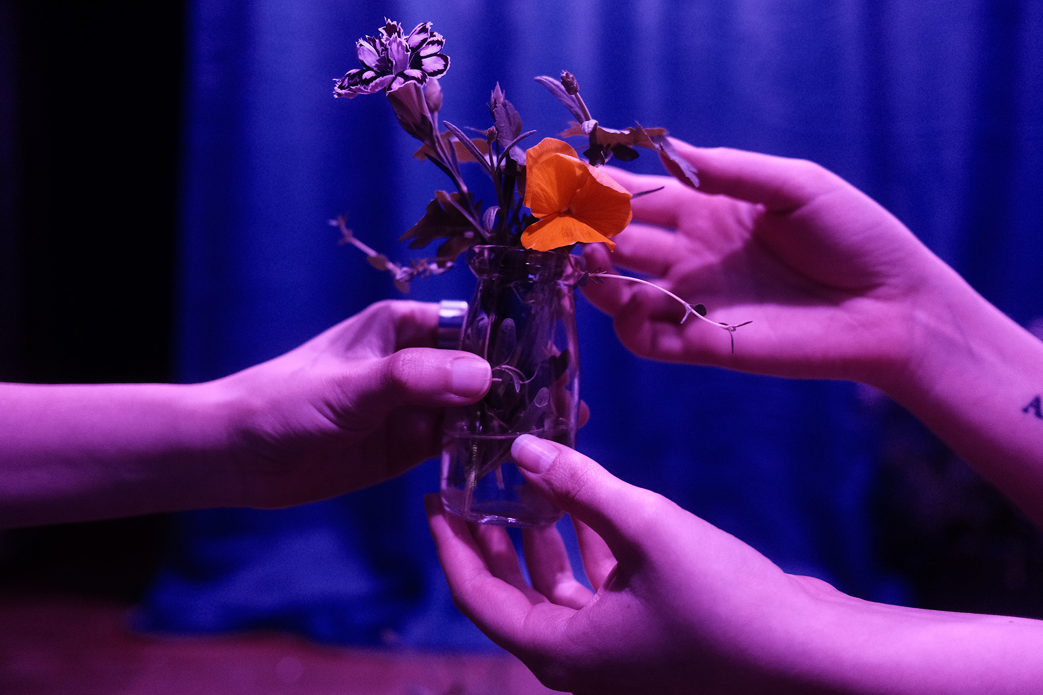 Image of Liz's thesis work. Photo shows the hands of two figures handing off a glass bottle filled with water, herbs, and flowers