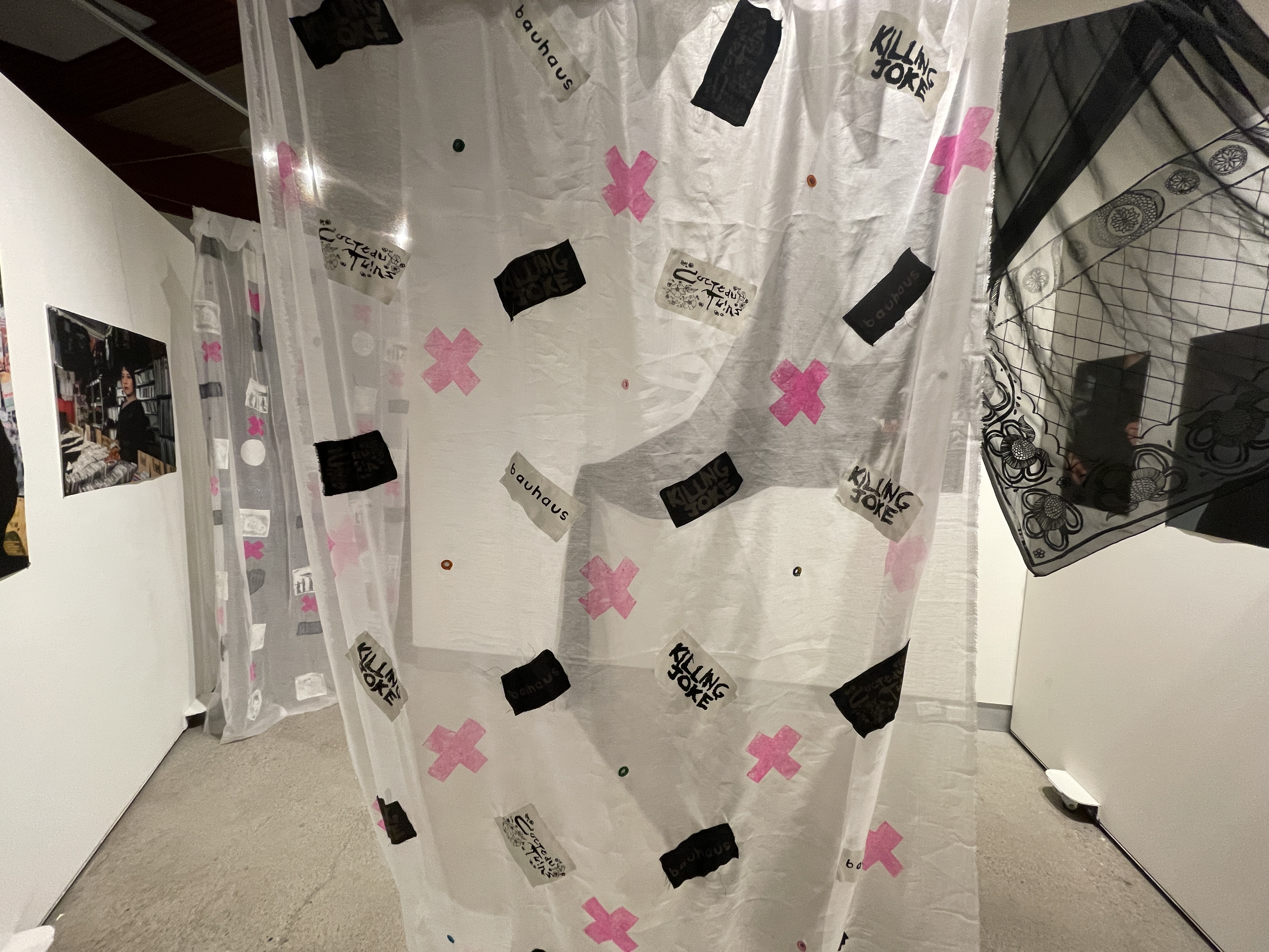 Image of Meher's thesis work, An interior view of an art installation featuring transparent curtains with various black and pink graphic elements, including text that reads 'Bauhaus' and 'KILLING JOKE'. The curtains are hung in a space with white walls, and are slightly overlapping, creating a partitioned area. On the left, a photograph is mounted on the wall, and the reflective surface on the right hints at the rest of the gallery space.