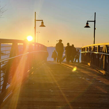 People are walking across a bridge on a snowy landscape and the sun is setting in the background.