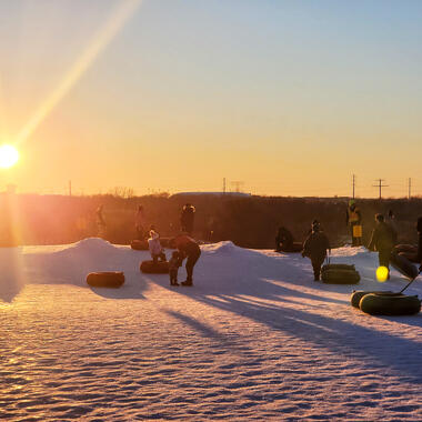 A snowy landscape at sunset, there are multiple people sledding and the sun is setting in the background