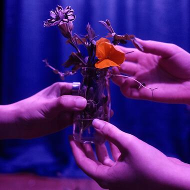 Image of Liz's thesis work. Photo shows the hands of two figures handing off a glass bottle filled with water, herbs, and flowers