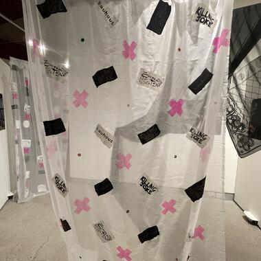 Image of Meher's thesis work, An interior view of an art installation featuring transparent curtains with various black and pink graphic elements, including text that reads 'Bauhaus' and 'KILLING JOKE'. The curtains are hung in a space with white walls, and are slightly overlapping, creating a partitioned area. On the left, a photograph is mounted on the wall, and the reflective surface on the right hints at the rest of the gallery space.