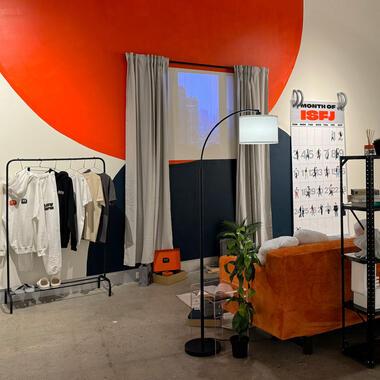 Image of Ria's thesis work. A modern room setup with a bold red accent wall. Clothes hang on a rack, a large floor lamp arcs over an orange couch, and a shelving unit holds various items, including books and electronics. A large window shows a cityscape, and a calendar titled 'MONTH OF ISFJ' is on the right.
