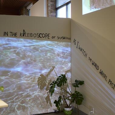 Image of Shafrin's thesis work. A corner of a room with walls displaying the projected text 'LOST IN THE KALEIDOSCOPE OF SENSATION' on one side and 'AS I WATCH THE WORLD WHIRL PAST ME' on the other. The projection casts ripples of light that resemble water over the walls and the shadow of a plant."