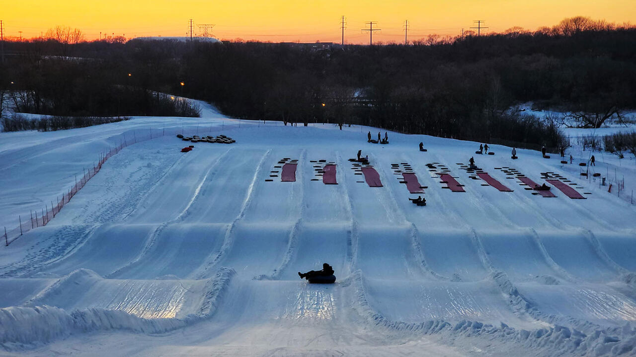 A landscape photograph of a sledding hill, there are several sledding lanes and the sun is setting in the background