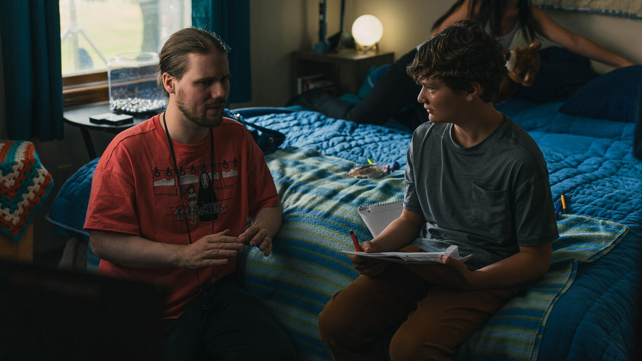 Two people sit on a bed and one of them is holding a notebook, the other is  a director giving them instructions