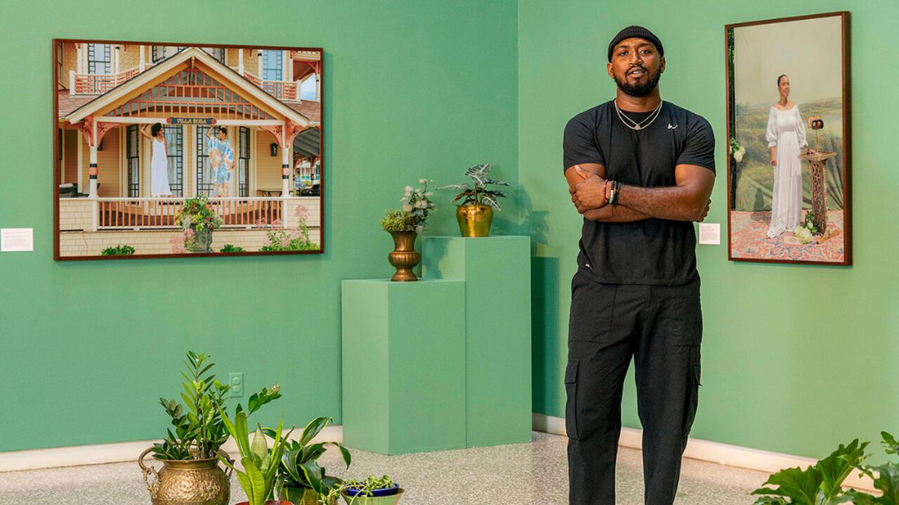 Bobby Rogers stands infront of a green wall, there are two paintings on the wall and several plants around the room