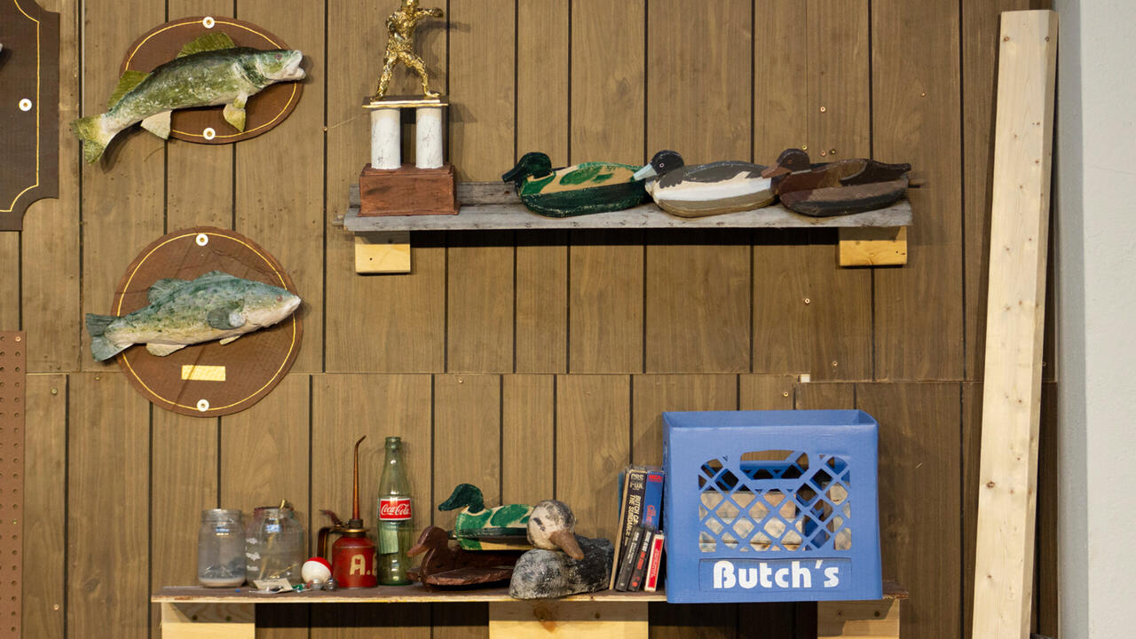 A wall of wooden panels, on it is a trophy, duck replicas, fish mounted on the wall, a soda bottle, and a bin of books