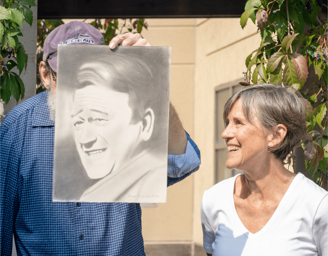An image of two people, one man holding up their drawing of a portrait, and a shorter woman looking at it.