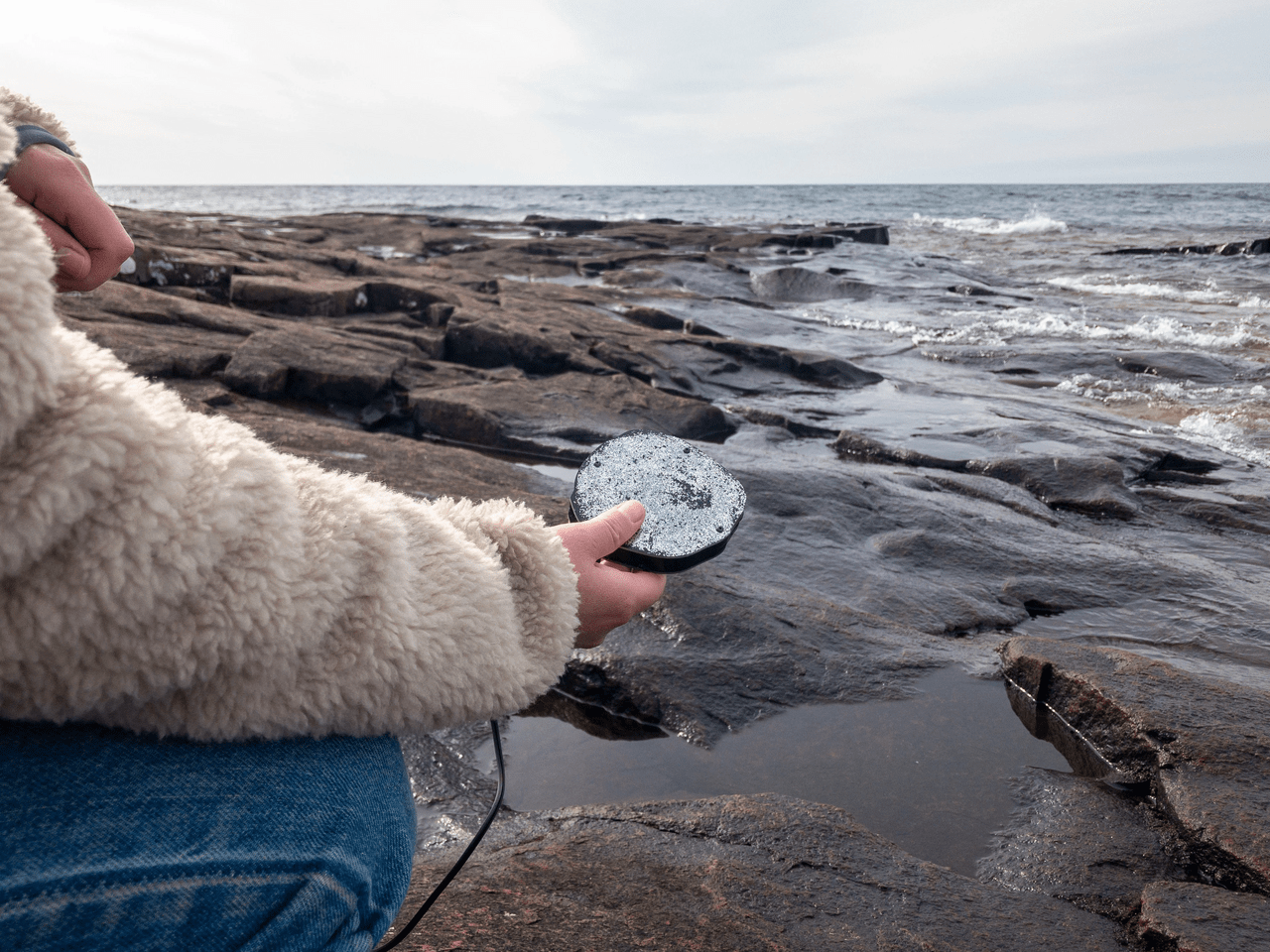 A photo of somebody holding a special flat rock in a shore scene full of larger, rougher rocks