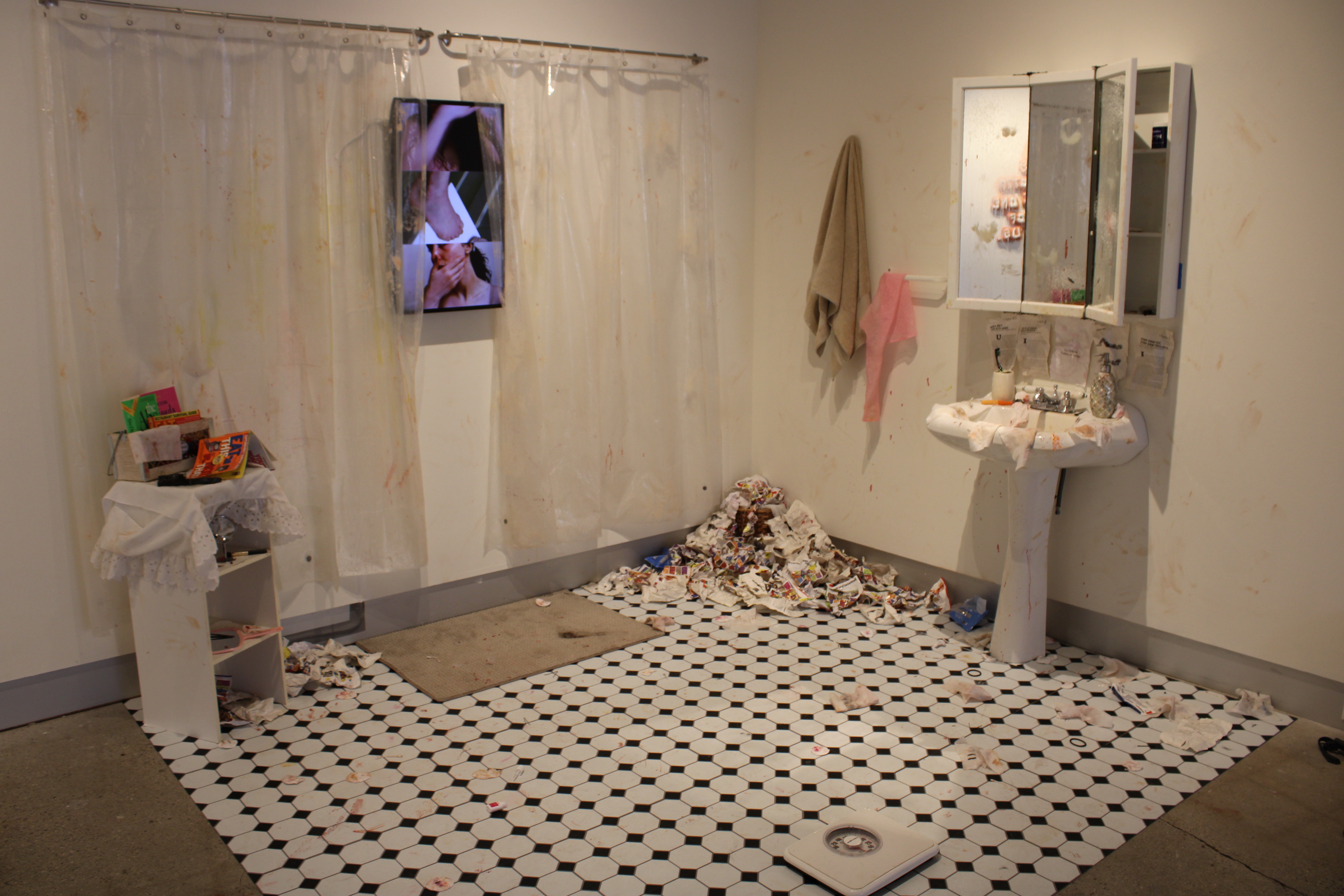 Artwork by Elsie Gray: an installation of a tiled bathroom floor, pedestal sink and mirror, overflowing wastebasket. A small television on the wall plays a video of the artist.