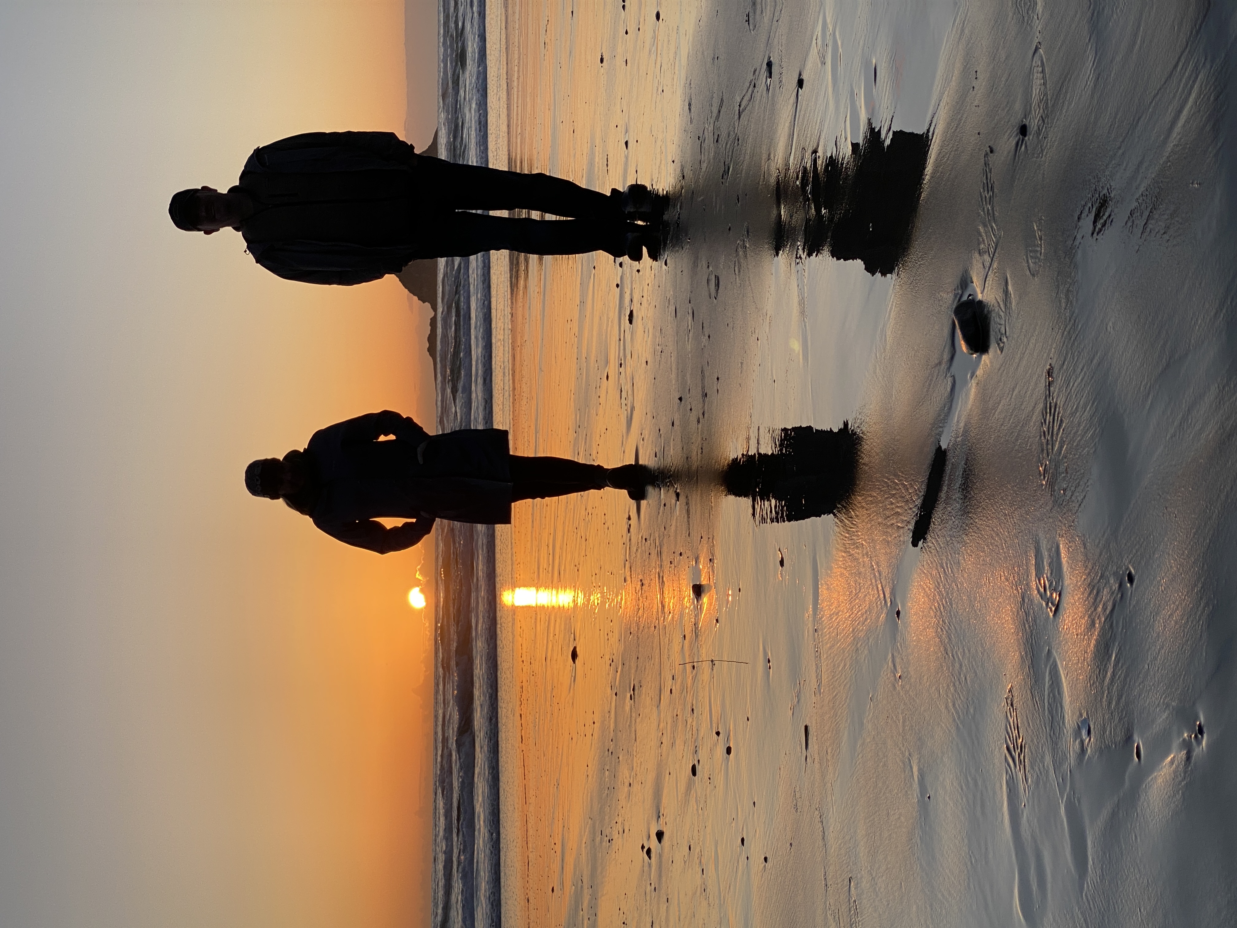 2 people standing side by side on a beach at sunset