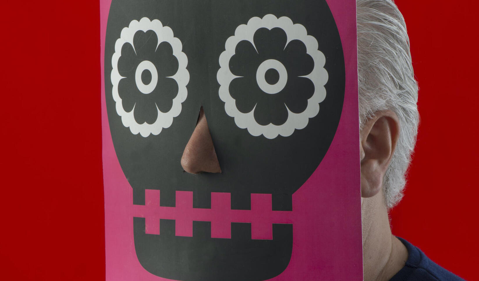 An older man holding his face up against a paper print of a decorative skull, and the nose is cut out ; Rik Sferra