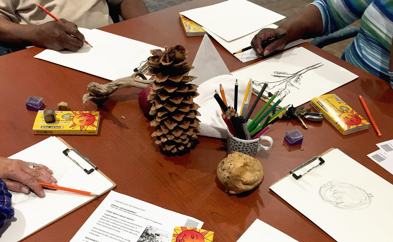 Drawing project with pineapple and drawing utensils in middle and students' hands on table