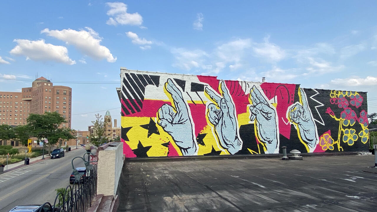 A brightly colored pink, white, and yellow mural on the side of a building featuring several hands making different hand shapes