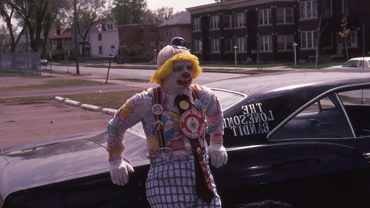 A photograph of a man dressed as a clown leaning up against a car