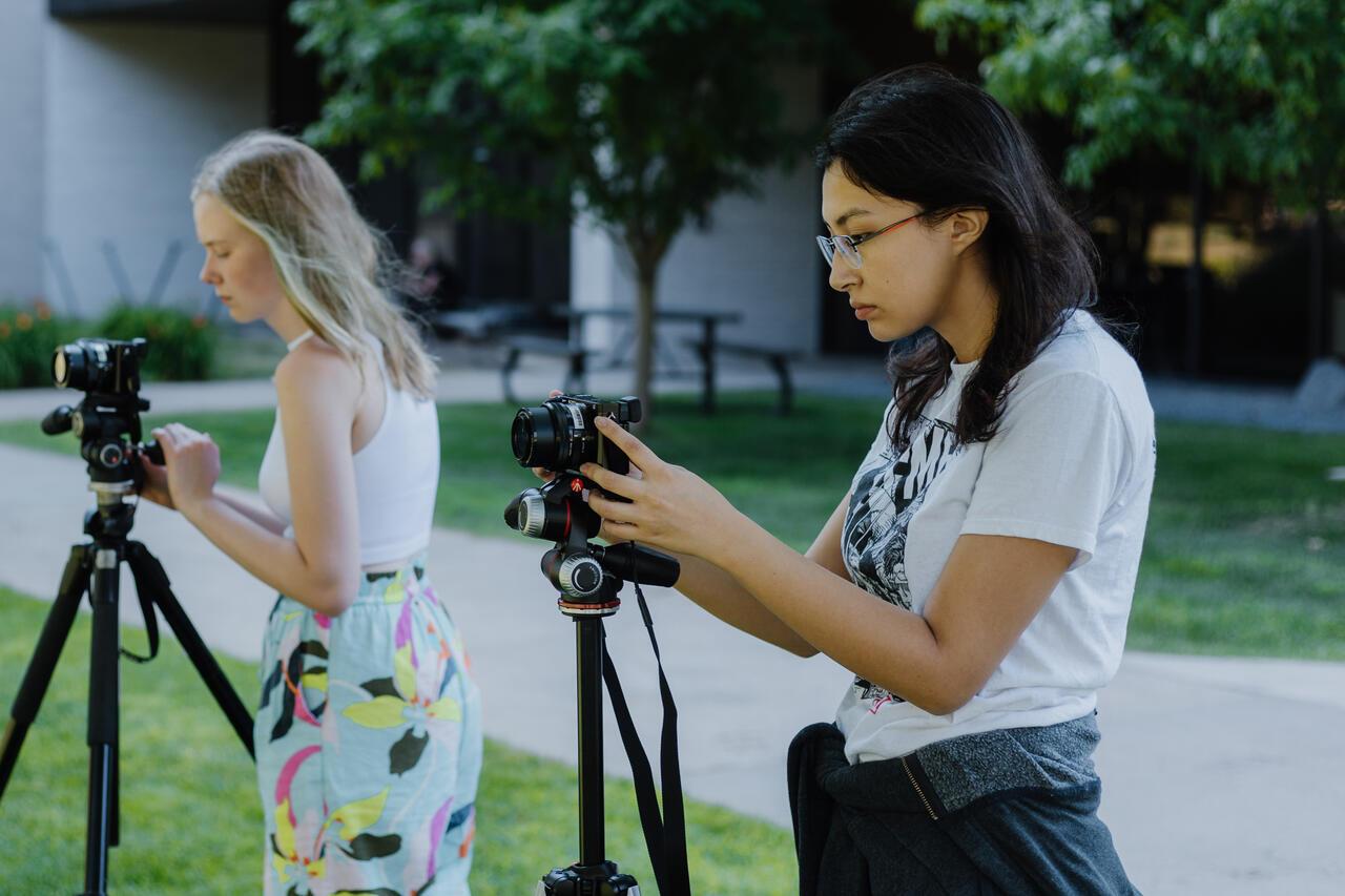 Two students stand outside with their cameras on tripods. 