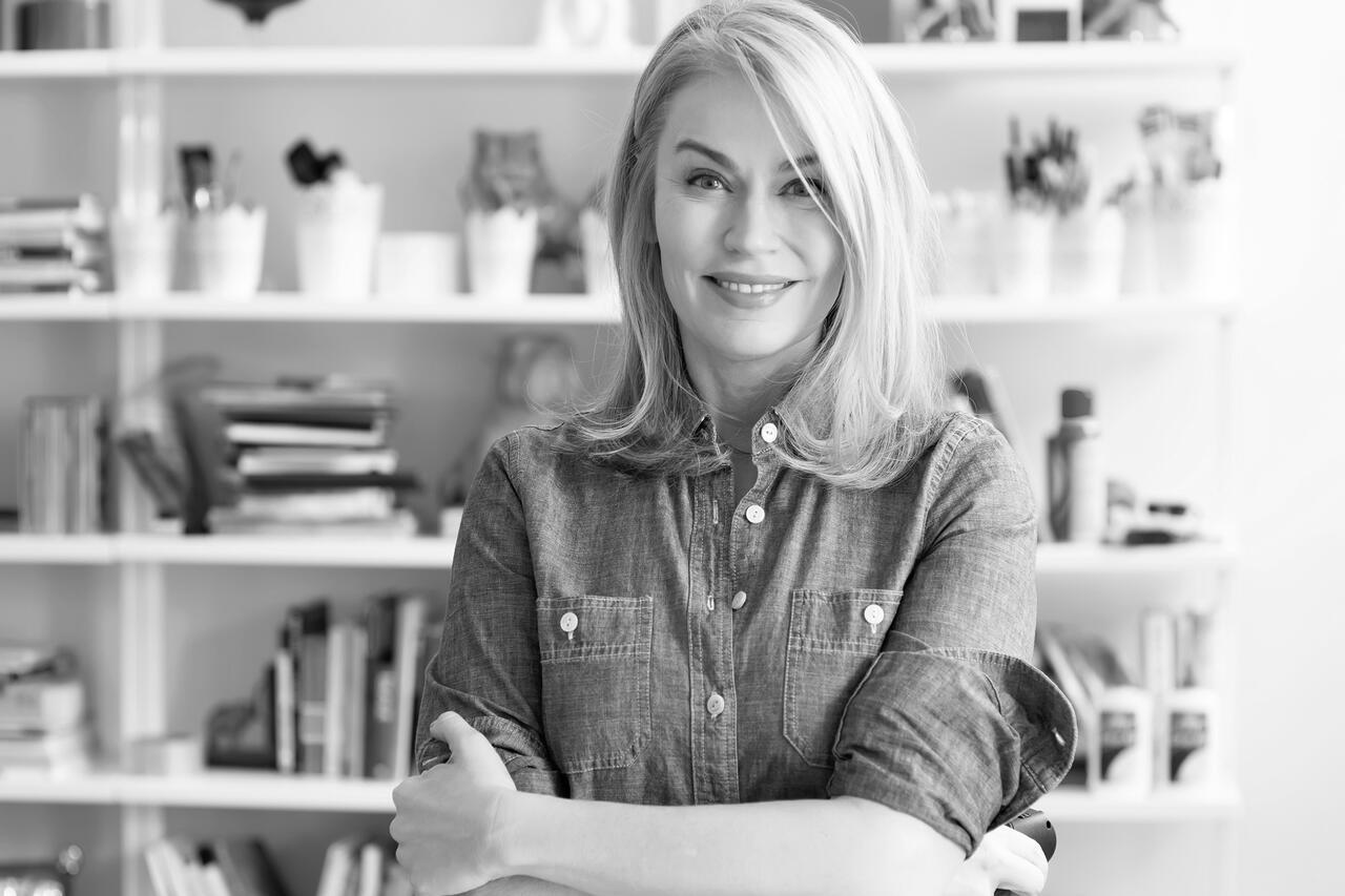 Black and white image of a woman with shoulder-length blonde hair wearing a button up shirt with her arms crossed. Behind her, you can see shelves of books and other supplies. Woman is looking directly into the camera and smiling.