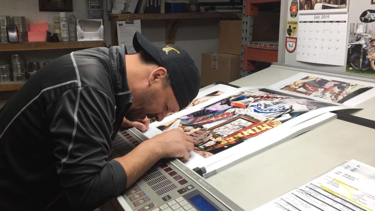 Tim Cortes bent over a drawing table working on an art piece featuring different hockey players, he is wearing a black hat and black jacket