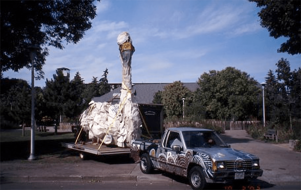 An image of a truck pulling a separate platform with a swan sculpture made of trash on it.