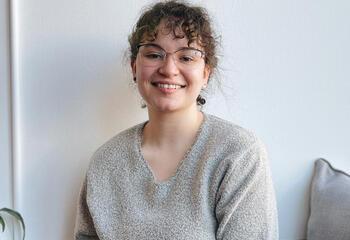 Portrait of Sarah Hormanski. She is sitting at a desk smiling with a gray shirt and jeans.